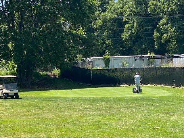 Greenskeeper Jimmy mowing a green at Silver Spring Golf Club in Riverside RI with a golf cart parked nearby
